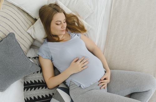 Беременная женщина лежит на кровати и держит живот двумя руками.