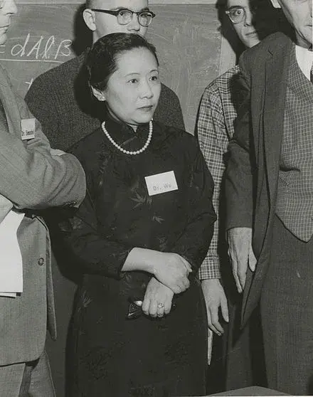 Black-and-white photograph of physicist Chien-Shiung Wu standing in a group of male colleagues. She wears a traditional Chinese dress with a pearl necklace and a name tag that reads “Dr. Wu,” highlighting her prominent role in mid-20th century physics, particularly her groundbreaking work on the Wu experiment.