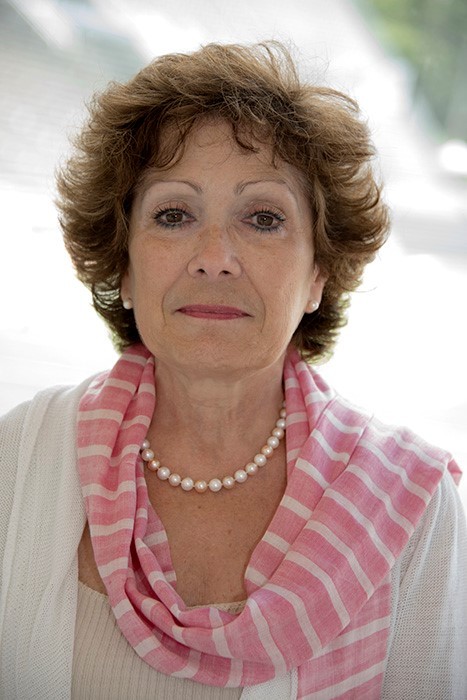 Portrait of Canadian neuroscientist Edith Hamel wearing a white outfit with a pink and white striped scarf and pearl necklace. She is standing outdoors, looking directly at the camera.