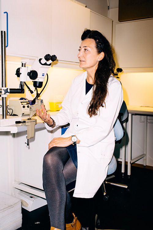 Norwegian neuroscientist May-Britt Moser wearing a white lab coat, seated at a lab bench beside a microscope. She is holding a small object, possibly a sample, and smiling slightly in a clinical research environment.