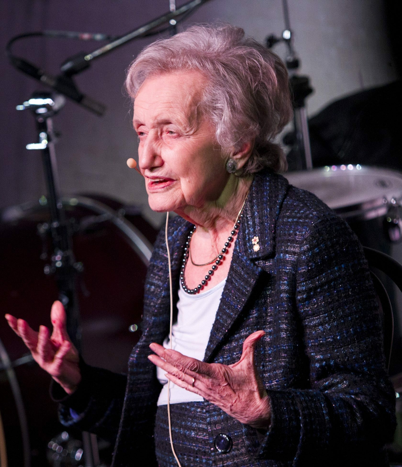 Brenda Milner, an elderly woman with short grey hair, speaks animatedly on stage while wearing a dark plaid jacket and a pearl necklace. She is using a headset microphone, and musical instruments like drums are visible in the background.