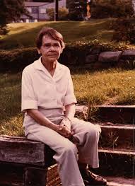 Sepia-toned photograph of Barbara McClintock, pioneering American scientist, sitting on wooden steps in a grassy outdoor area. She is wearing a short-sleeved buttoned shirt and trousers, with a calm and contemplative expression.
