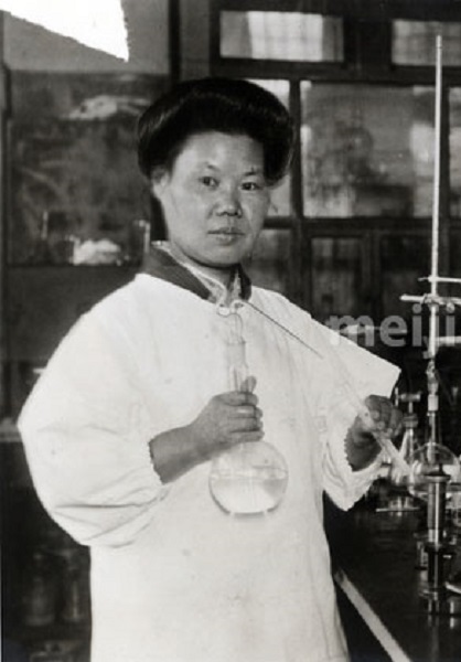 Historic black-and-white photograph of Japanese biochemist Michiyo Tsujimura in a science lab, wearing a white lab coat and holding a round-bottom flask, known for discovering Vitamin C in green tea in 1924.
