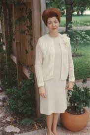 Historic photograph of Bette Nesmith Graham, inventor of Liquid Paper, standing outdoors in a garden setting. She is wearing a cream-colored dress and cardigan, with a floral corsage on her left shoulder, and has styled red hair. A potted plant and wooden trellis are visible behind her.