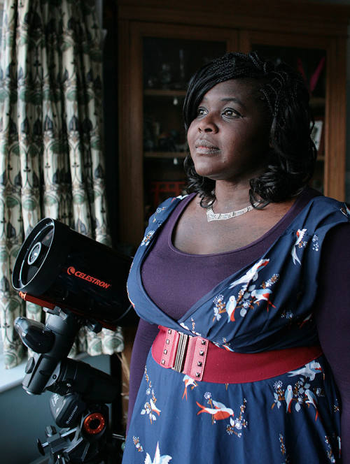Maggie Aderin-Pocock, British space scientist and science communicator, smiling and standing next to a black Celestron telescope in a warmly lit room.