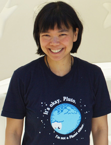 A smiling Jane X. Luu, a Vietnamese-American astronomer, wearing a navy blue t-shirt that features a cartoon drawing of Pluto with the words “It’s okay, Pluto. I’m not a planet either.”