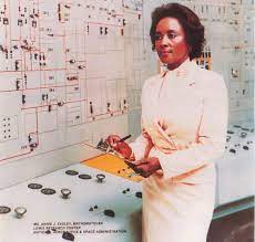 Historic photograph of a Black woman in a light-colored dress standing in front of a complex control panel, holding a clipboard and pen. She is Dr. Annie Easley, a NASA scientist and mathematician, known for her work in rocket propulsion and computer programming