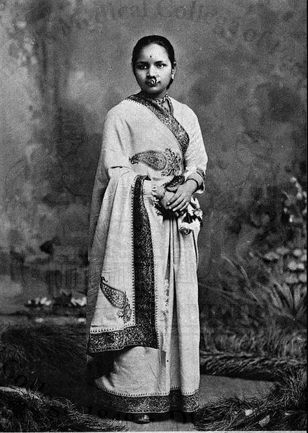 Historic black and white portrait of Anandibai Joshi, India’s first female doctor, standing in traditional attire with ornate embroidery, wearing a nose ring and jewelry, in a studio setting with a textured backdrop.
