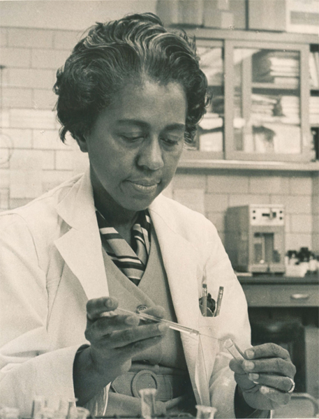 A black-and-white photograph of Marie Maynard Daly, an African-American biochemist, in a lab coat working with scientific glassware in a laboratory. She appears focused and calm as she conducts an experiment.