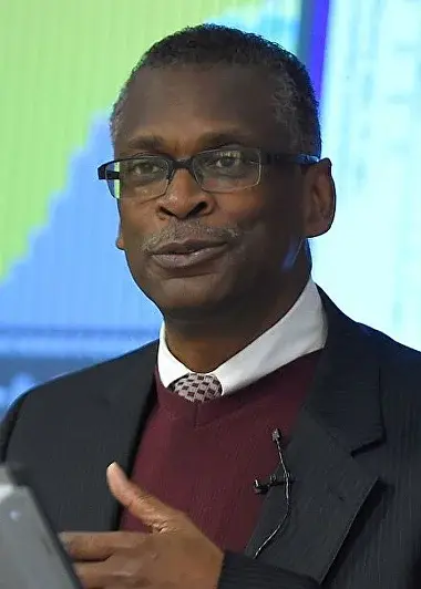 African-American inventor Lonnie G. Johnson speaking at a public event. He is wearing glasses, a burgundy sweater, and a suit jacket, with a microphone clipped to his collar. Behind him, colorful data graphs are visible on a screen.