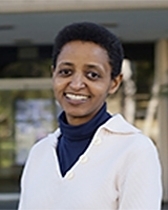 Historic black and white portrait of Anandibai Joshi, India’s first female doctor, standing in traditional attire with ornate embroidery, wearing a nose ring and jewelry, in a studio setting with a textured backdrop.