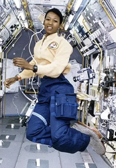 Mae Jemison floating in zero gravity aboard a spacecraft. She is wearing a yellow NASA flight suit with blue cargo trousers and smiling, surrounded by scientific equipment and control panels.